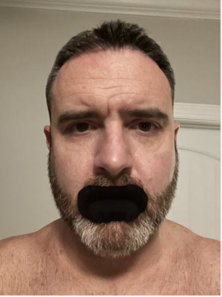 Man with a beard and a black mouth cover, standing indoors.