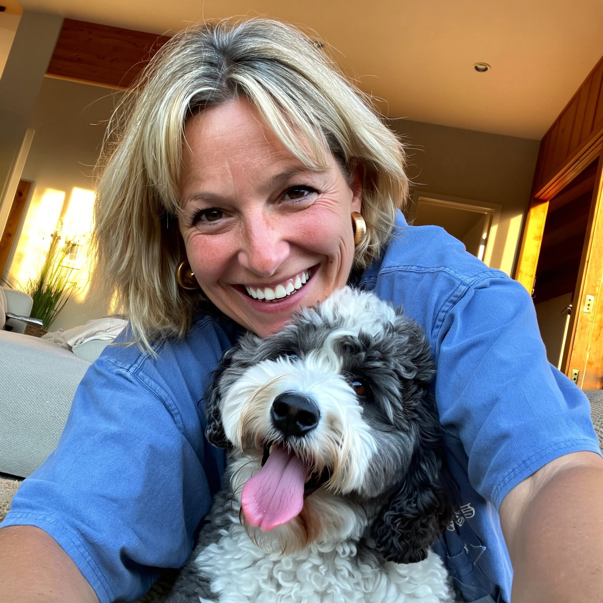 A smiling blonde woman in a blue shirt takes a selfie with her fluffy black and white dog.