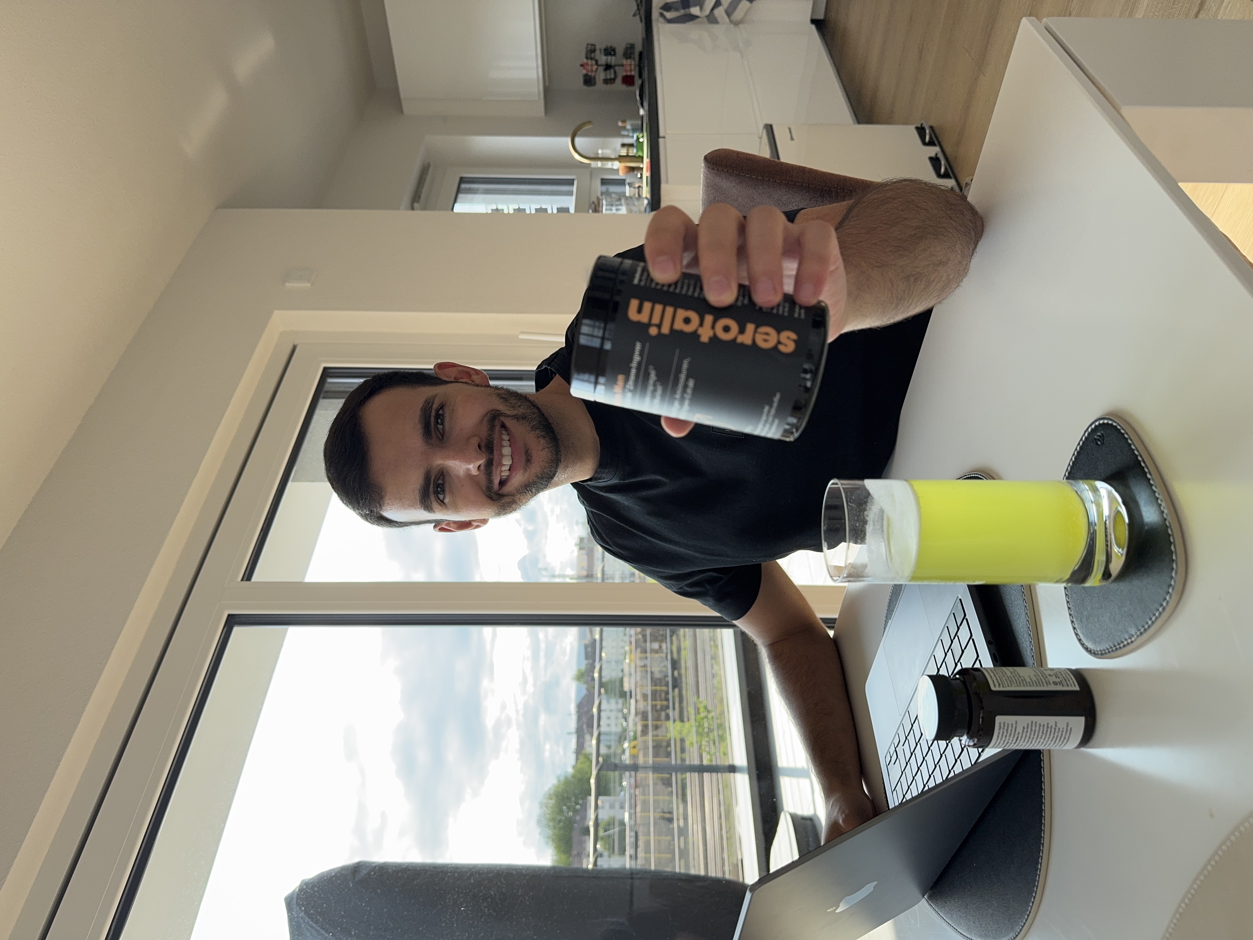 Person smiling, holding a container, sitting at a table with a laptop and a green drink.
