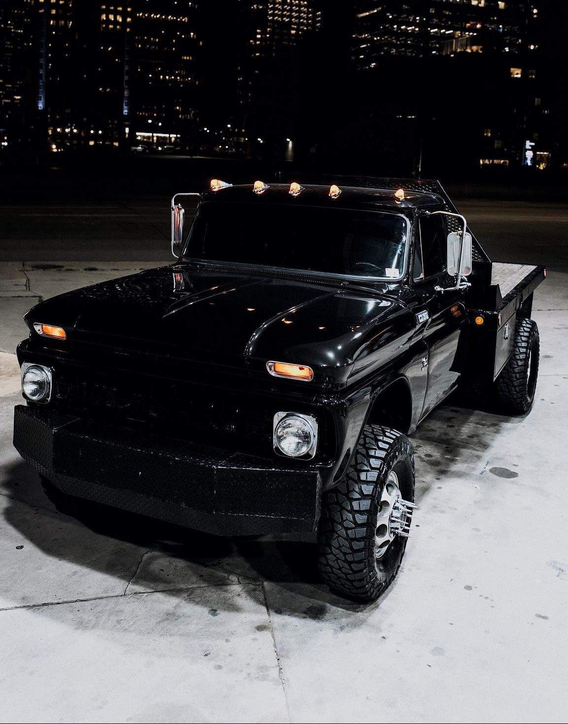 A customized black vintage flatbed truck parked at night with a city skyline in the background.