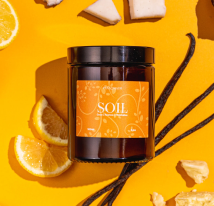 Brown candle jar labeled 'SOIL' with orange slices and vanilla pods on a yellow background.