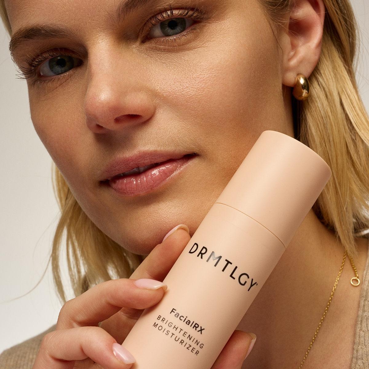 A close-up of a woman holding a bottle of DRMTLGY FacialRx Brightening Moisturizer near her face.