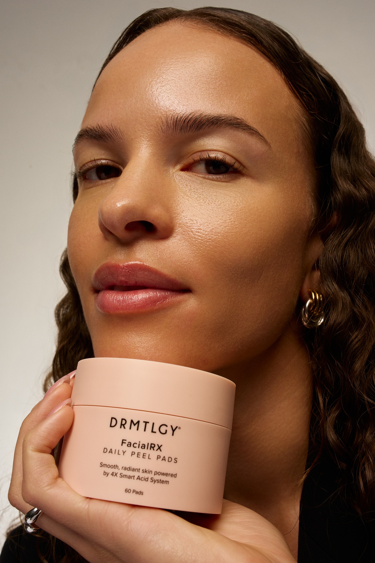 A woman with radiant skin holds a pink container of DRMTLGY FacialRX Daily Peel Pads.