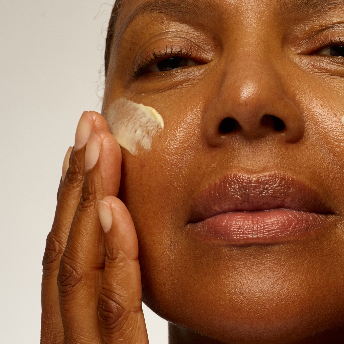 A close-up shot of a person with dark skin applying cream to their cheek with their fingers.