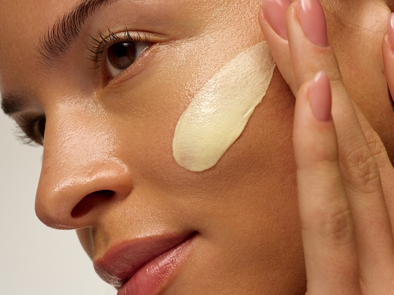 A close-up of a person with glowing skin applying a creamy, yellow product to their cheek.