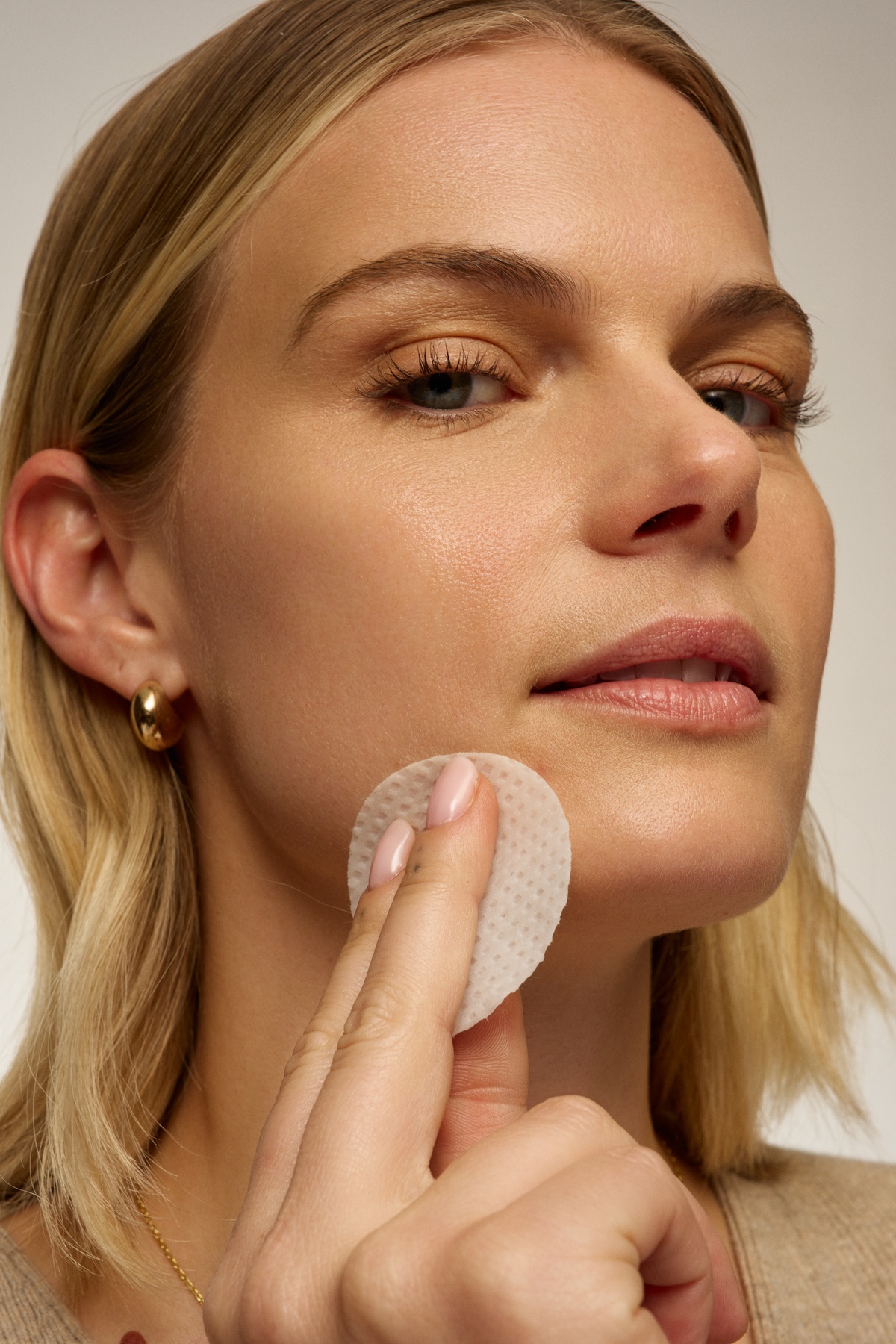 A close-up of a blonde woman holding a round, white, textured facial pad to her cheek.