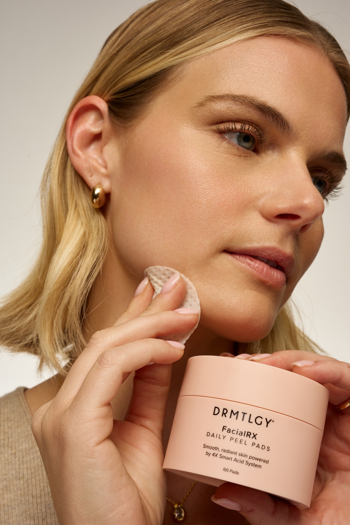 A woman applies a DRMTLGY Facial RX Daily Peel Pad to her cheek while holding the container.