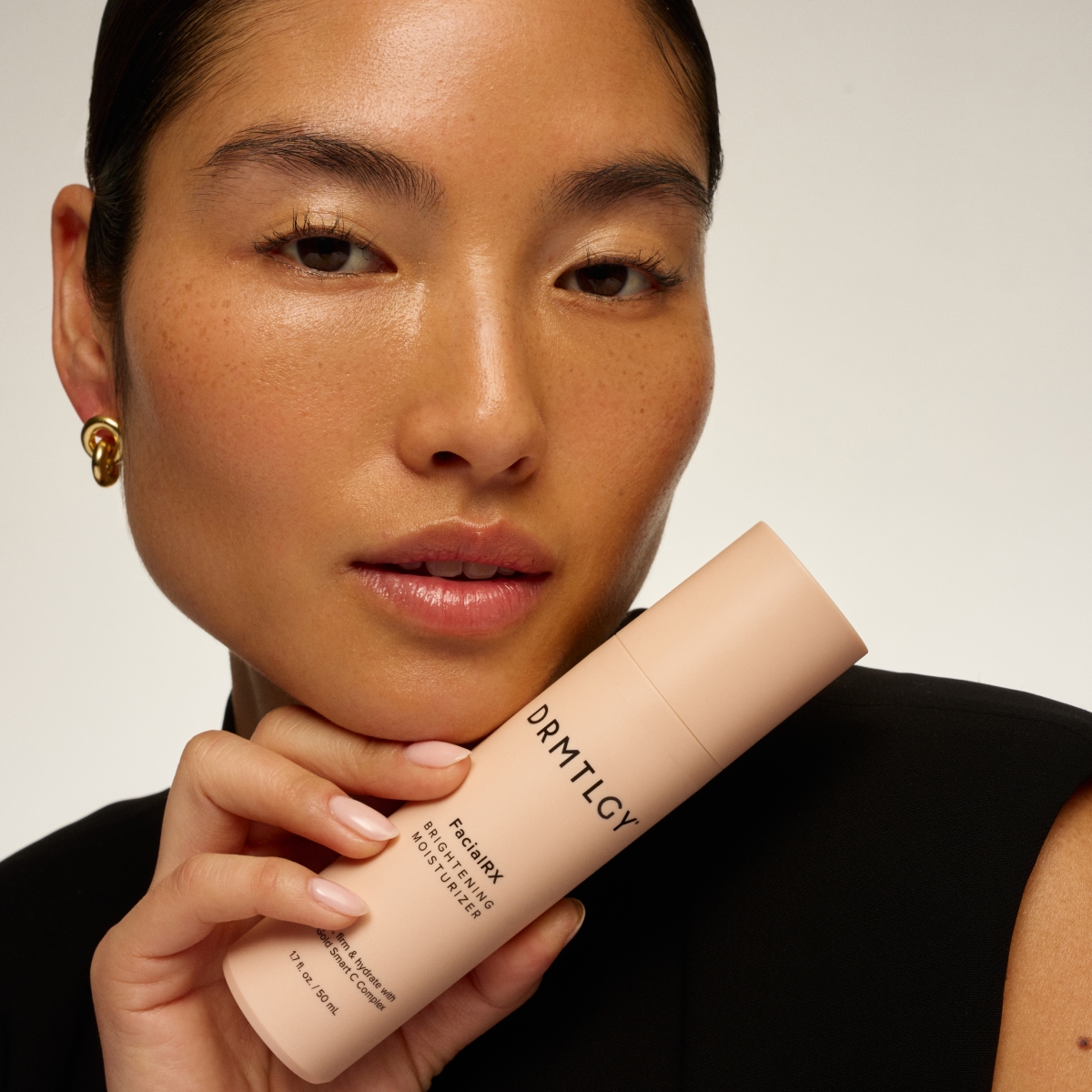 A woman with glowing skin holds a bottle of DRMTLGY Brightening Moisturizer against her chin.