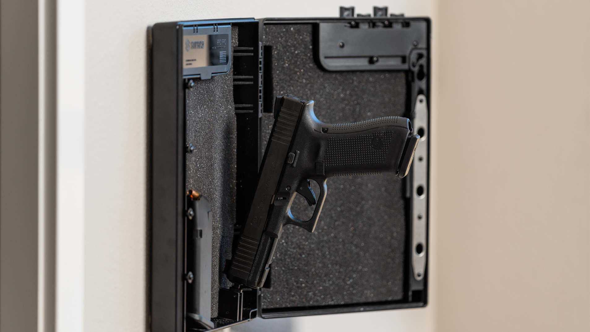 A black handgun and a magazine resting inside an open, wall-mounted gun safe with foam padding.
