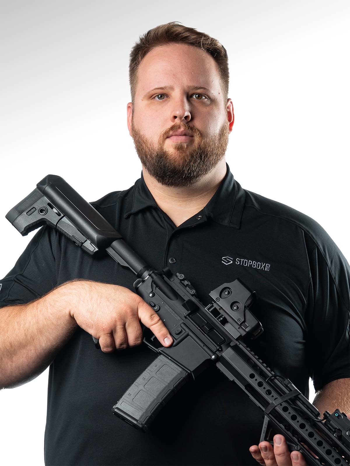 Man holding a rifle against a white background, wearing a black polo shirt.