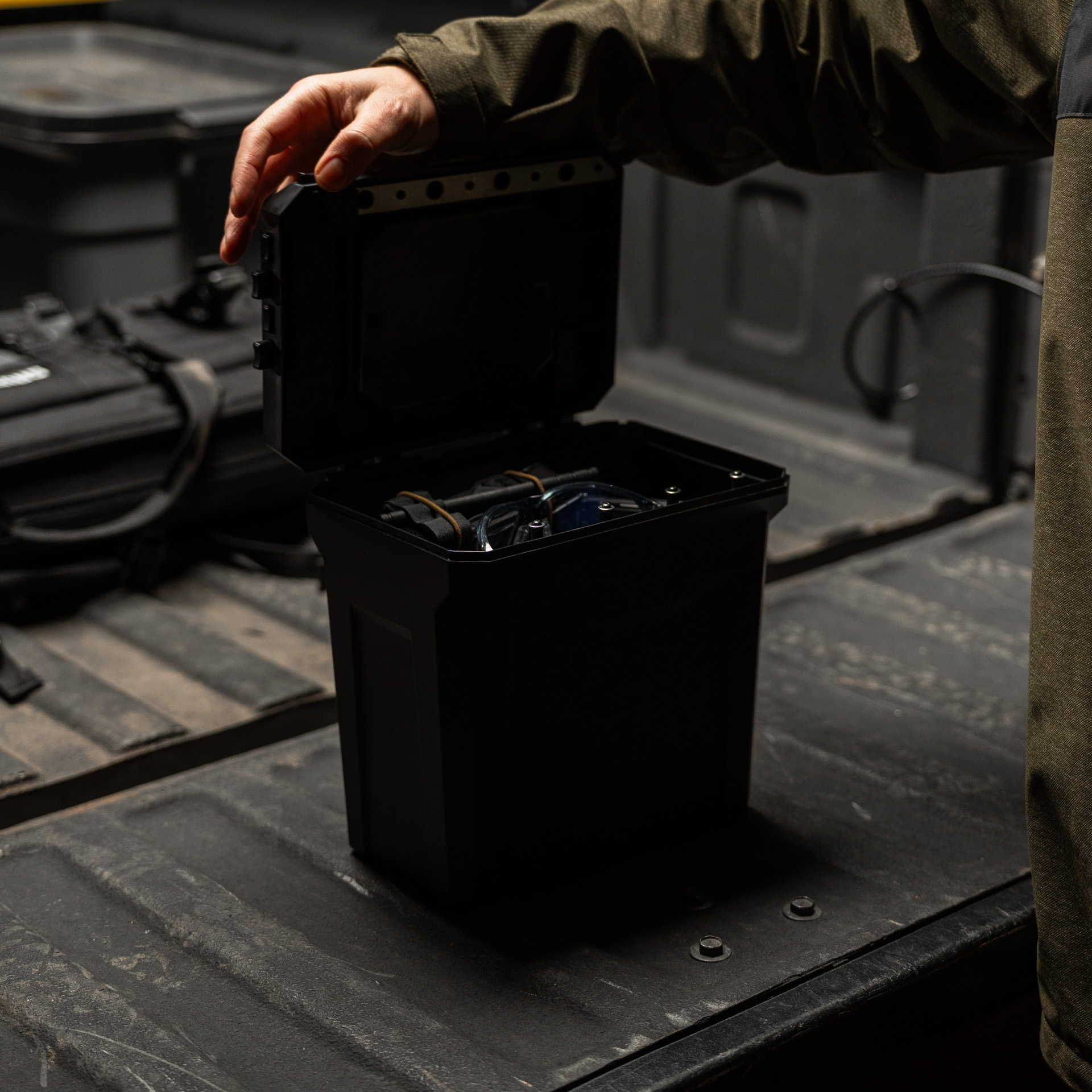 A person in a green jacket opens a black storage box in the bed of a truck.