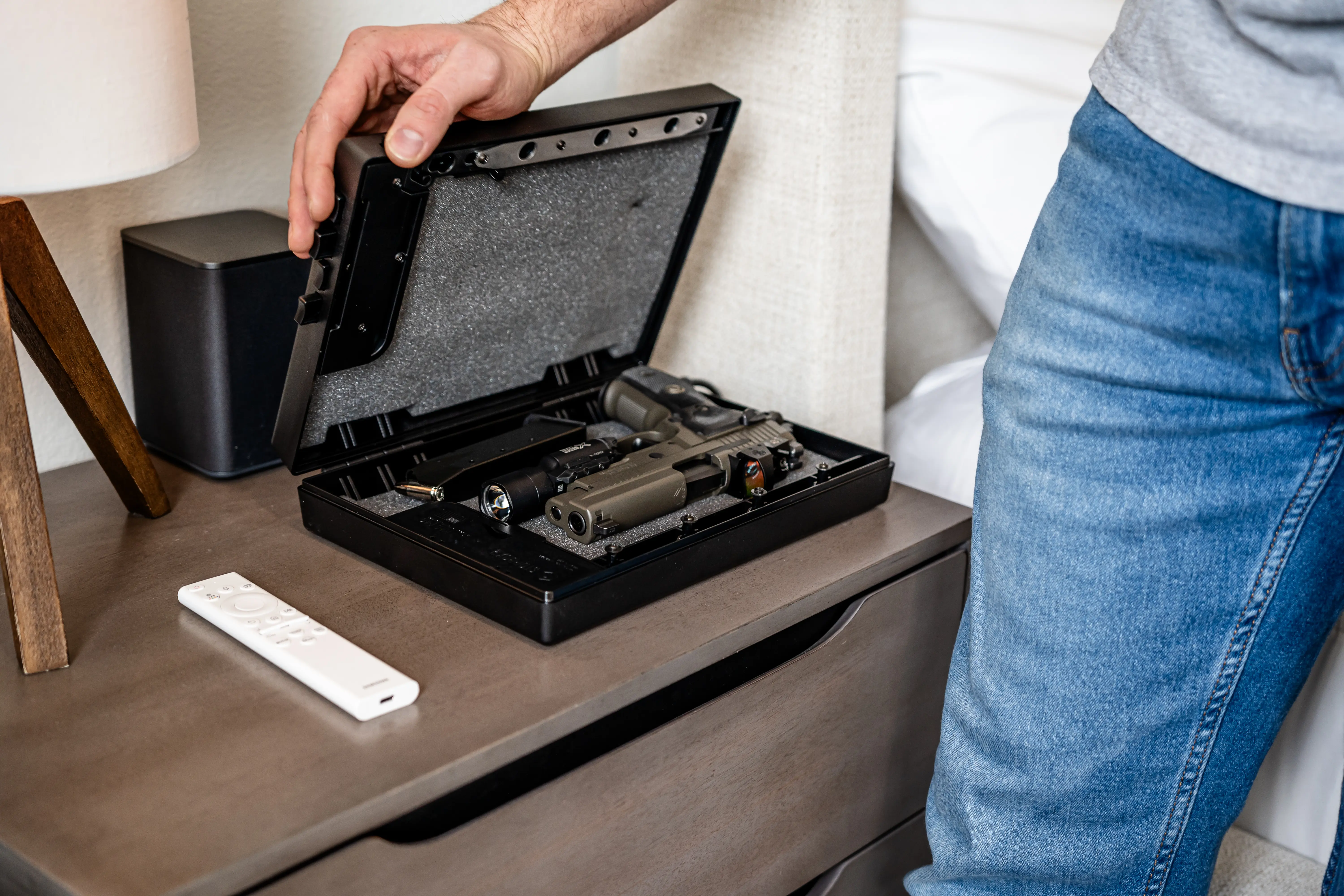 Person opening a gun safe on a bedside table with a remote control nearby.