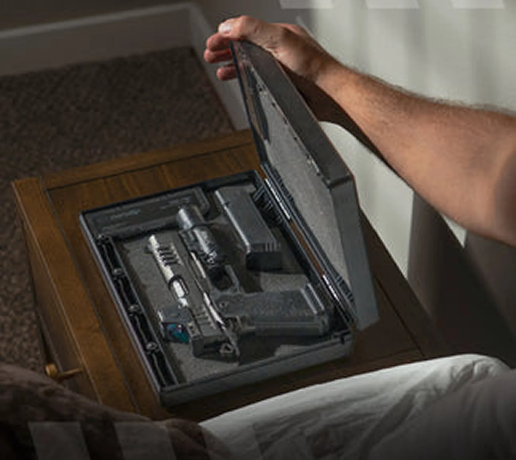 A person opens a small gun safe on a nightstand, revealing a handgun and magazine inside.