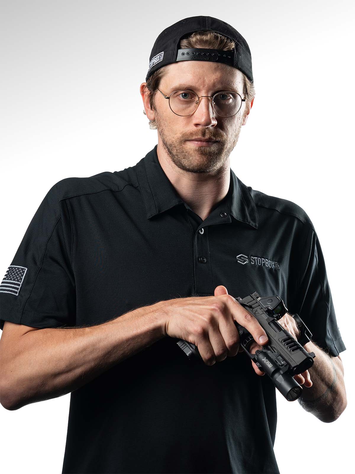 Man holding a firearm, wearing glasses and black cap, against white background.
