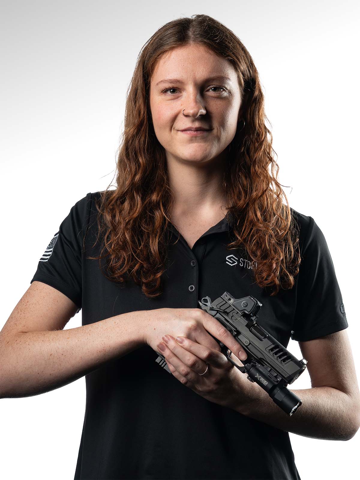 Person with long hair holding a firearm, wearing a black polo shirt.