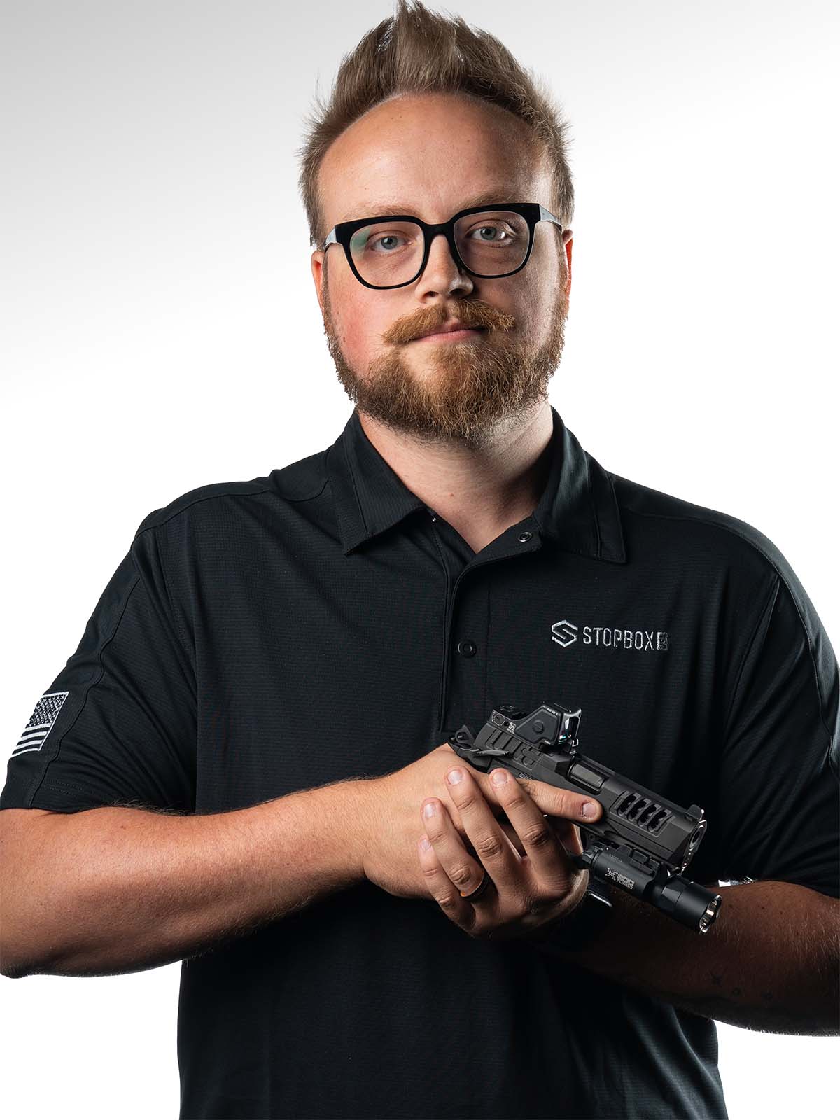 Person wearing glasses and black shirt holding a firearm against a white background.