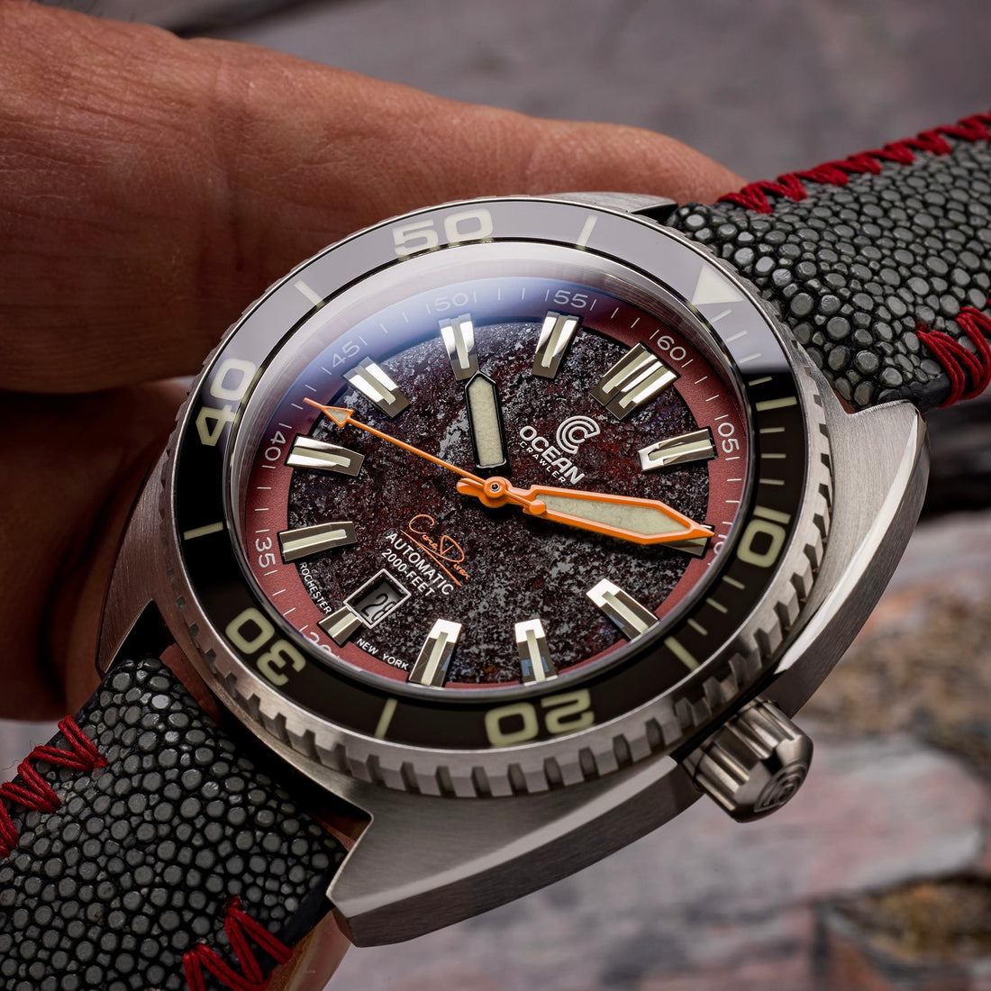 A close-up of a watch with a textured, reddish-brown face and a black, pebbled strap with red stitching.