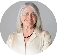 Headshot of a smiling woman with long, straight gray hair, wearing a cream blouse and a dark necklace.
