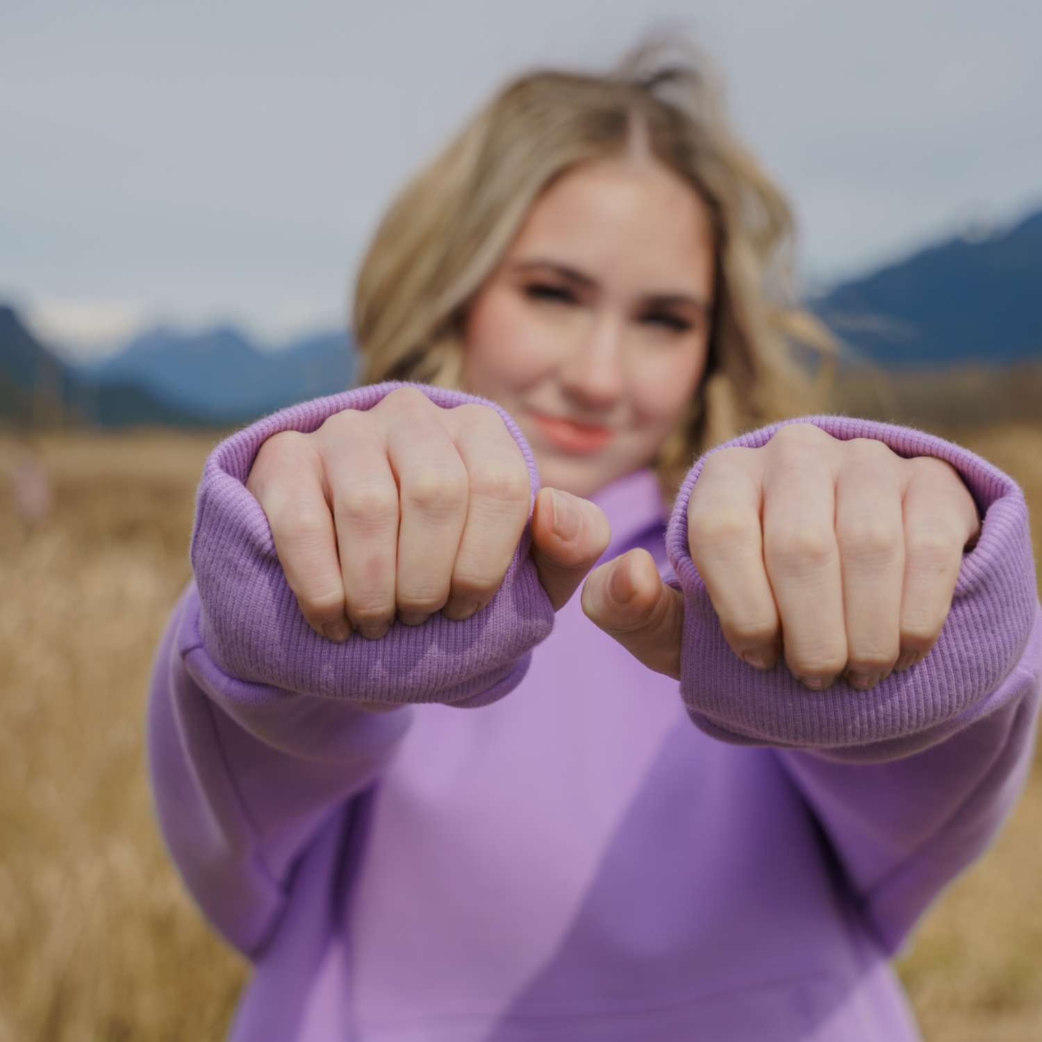 Person in purple hoodie showing fists outdoors with mountains in background.
