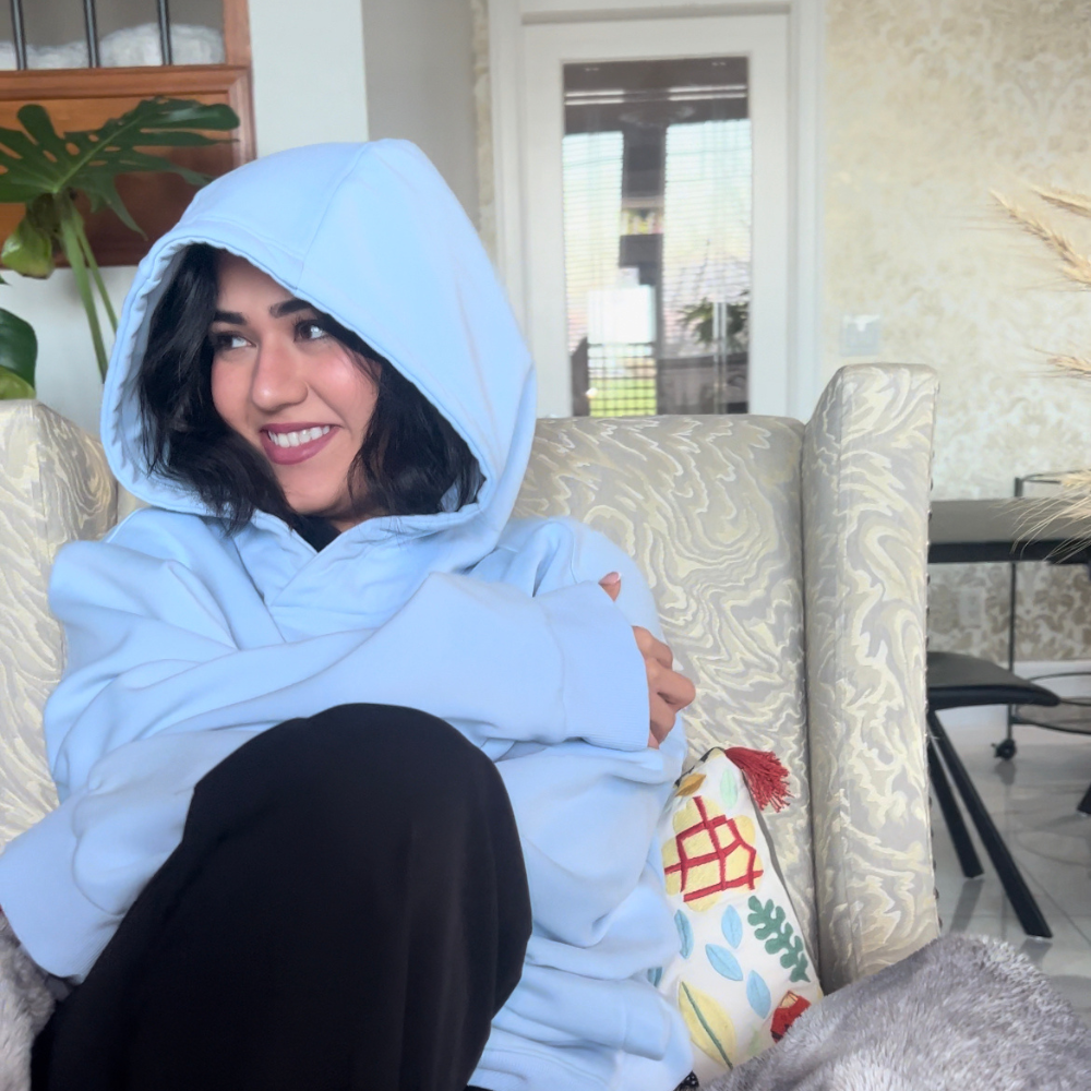 Person in a blue hoodie sitting on a patterned chair, smiling.