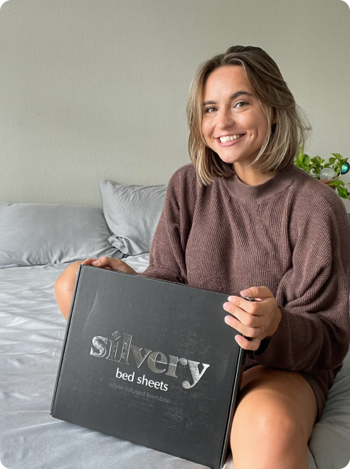 Person holding a box of silvery bamboo bed sheets, sitting on a bed.