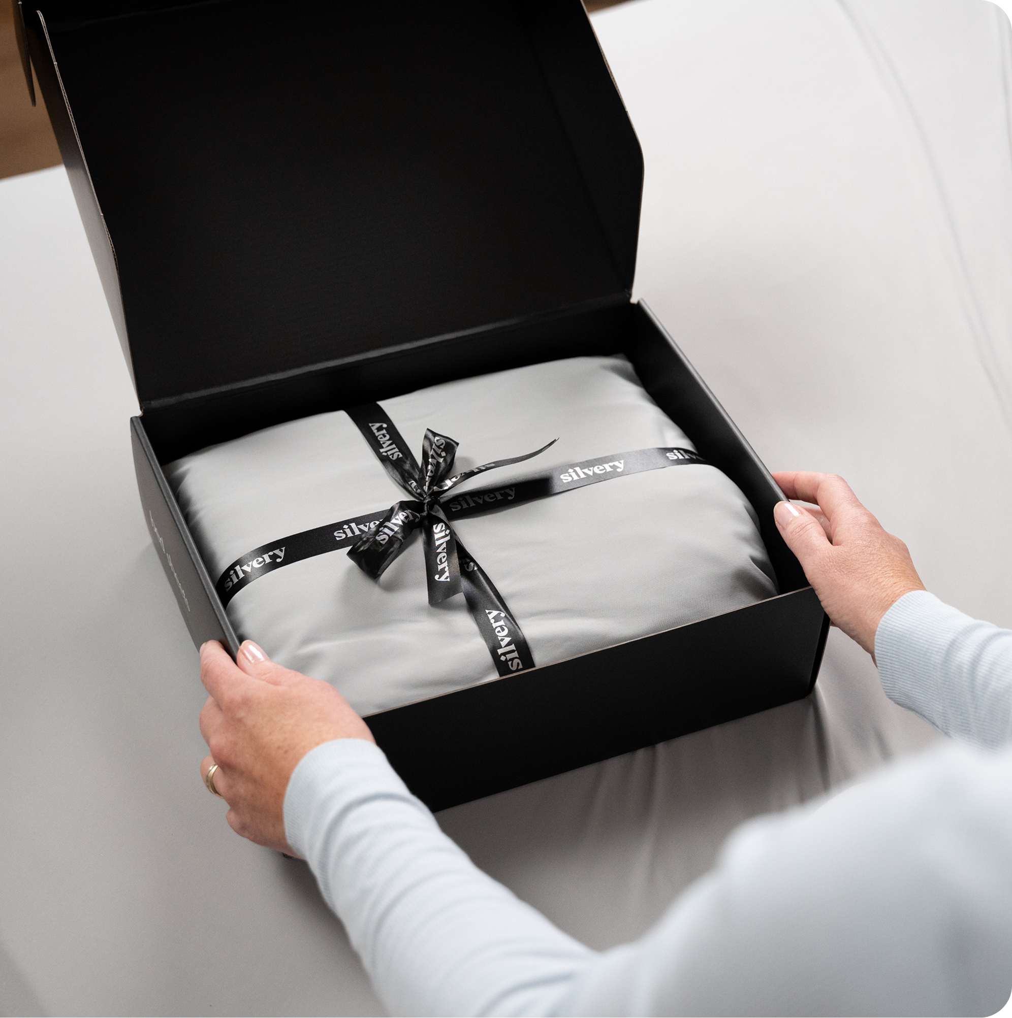 A person's hands opening a black gift box containing a grey fabric item tied with a branded ribbon.