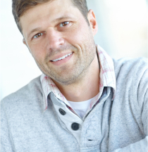 A man smiling, wearing a gray sweater with a plaid shirt underneath.