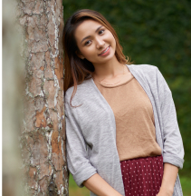 Person smiling, leaning against a tree, wearing a gray cardigan and brown top.