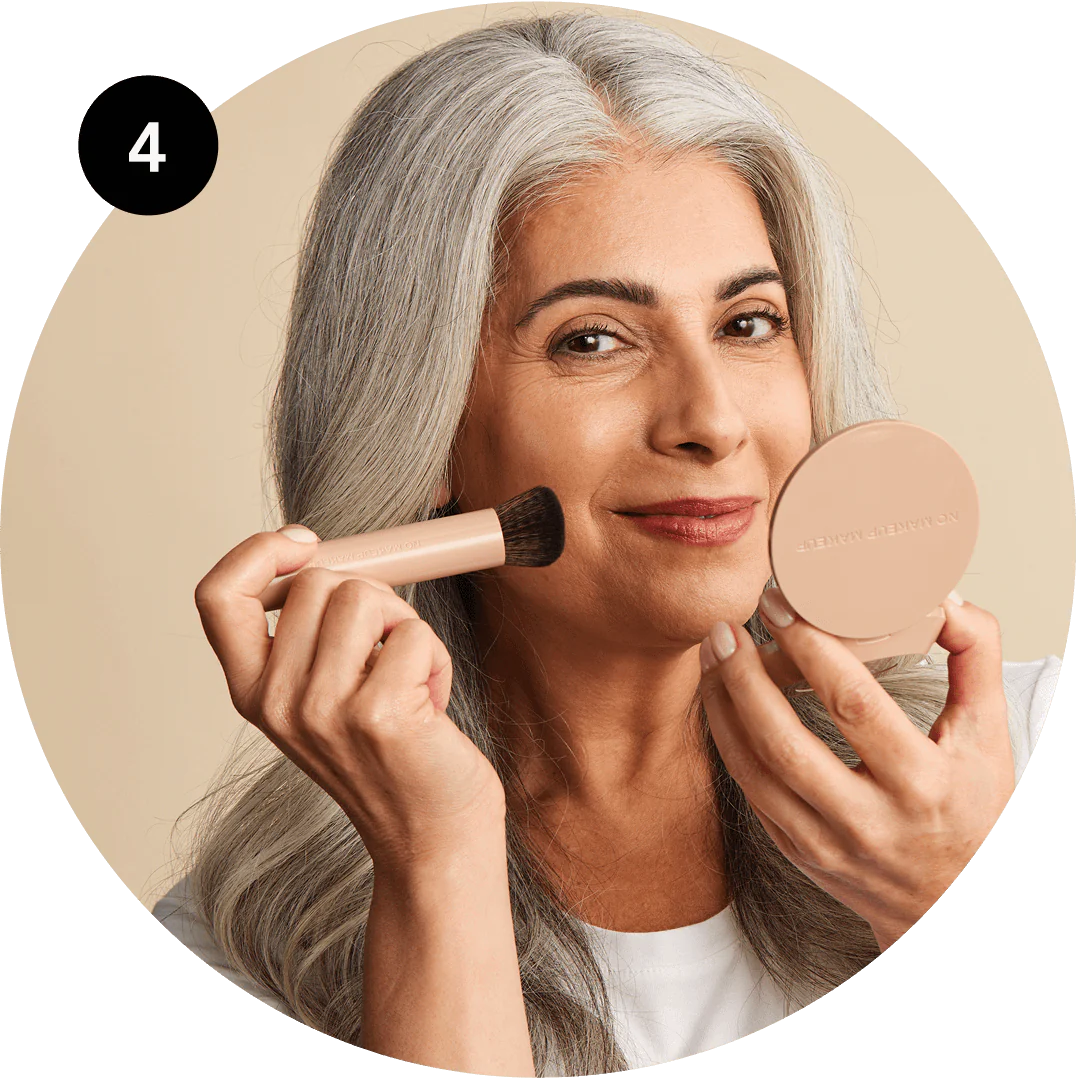 Woman with gray hair applying makeup with a brush and compact mirror.