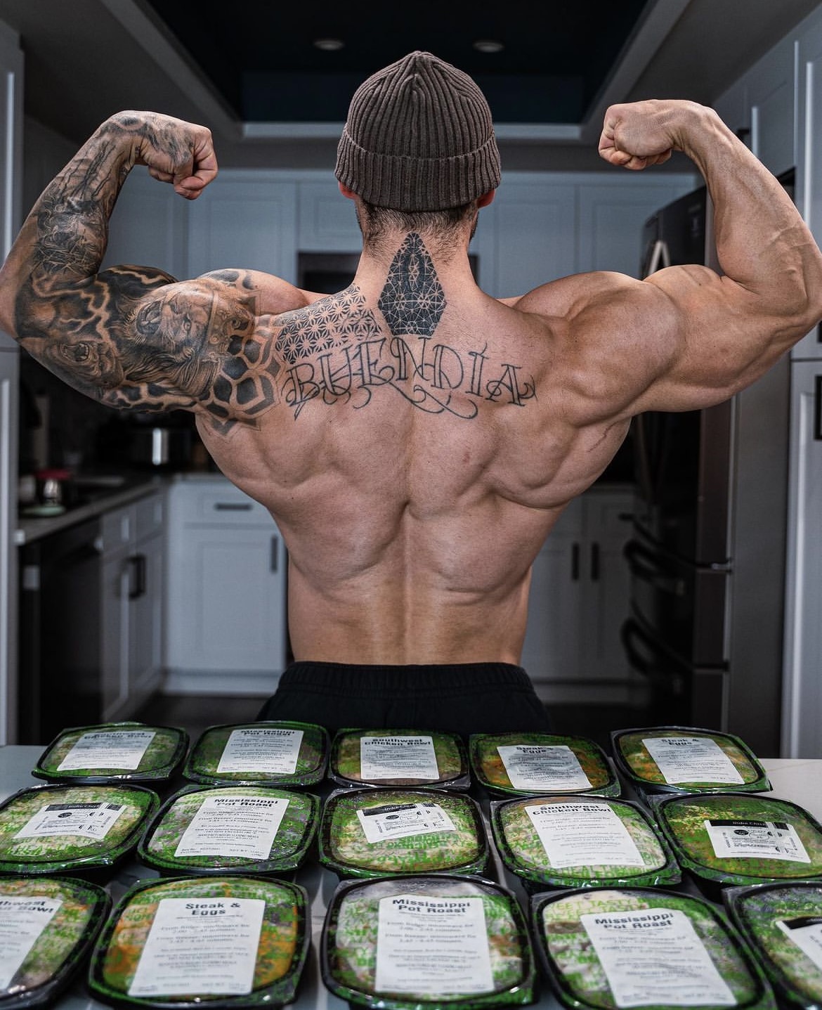 Person flexing back muscles with tattoos, standing in kitchen with meal containers on counter.