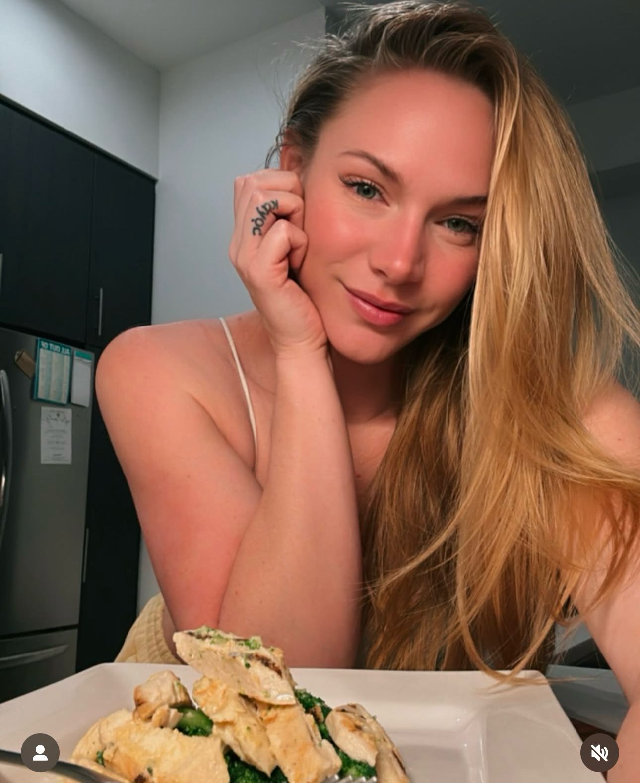 Person with long hair posing with a plate of food in a kitchen.