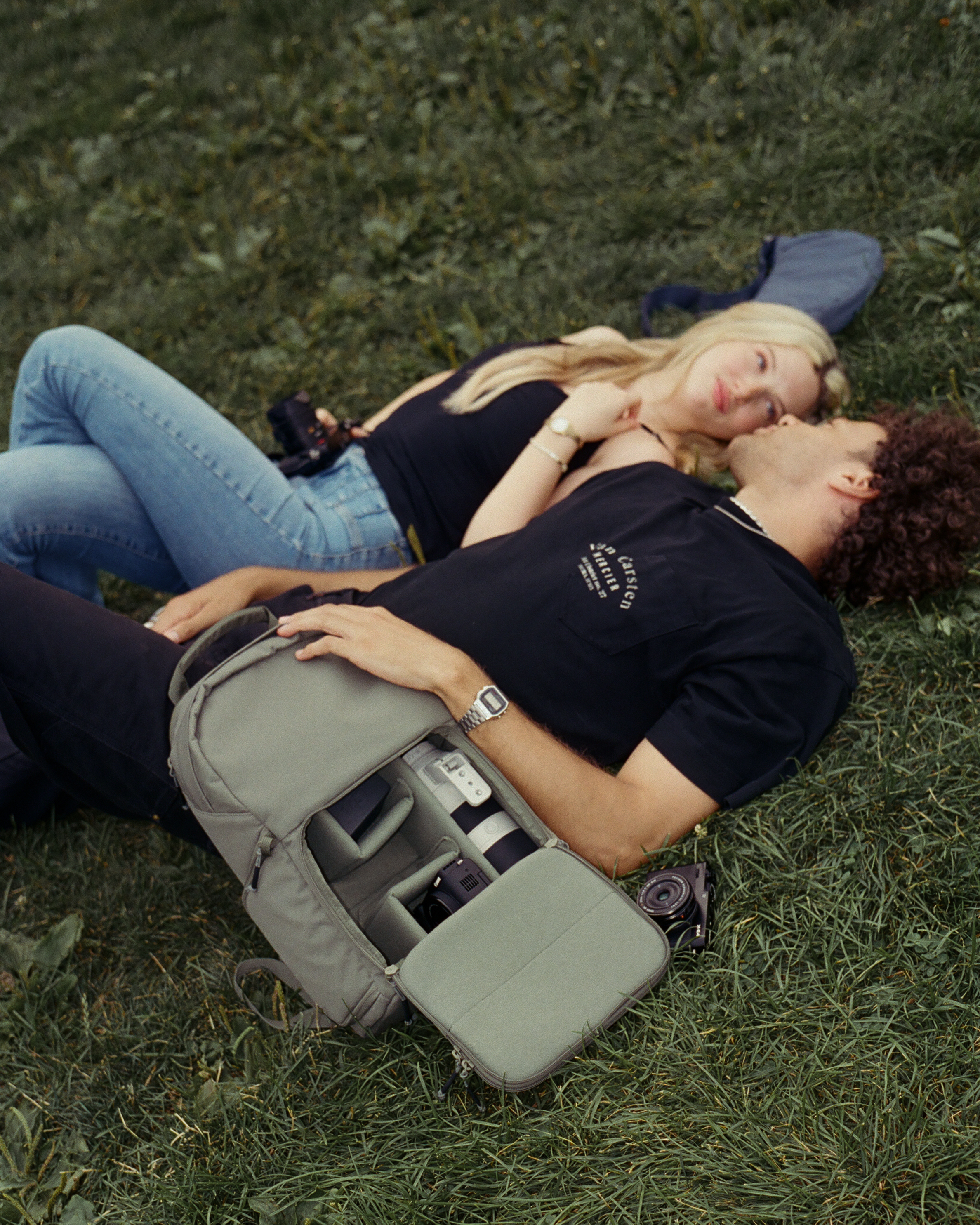Two people lying on grass with a camera bag and cameras.