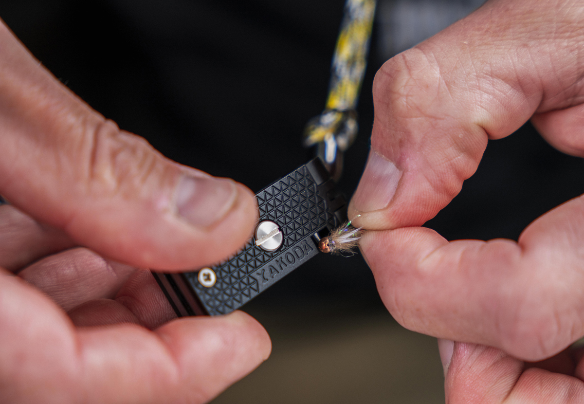 Hands holding a small black tool and a fishing fly.