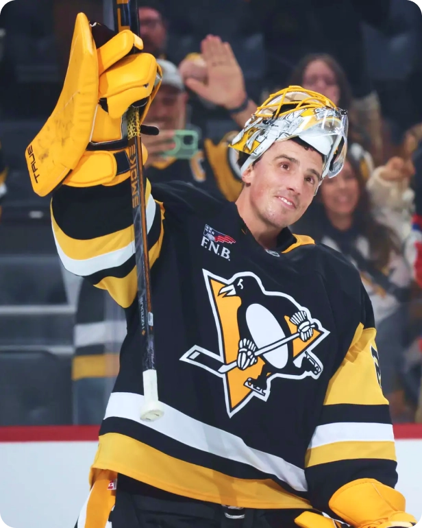 A smiling Pittsburgh Penguins hockey goalie raises his stick on the ice.