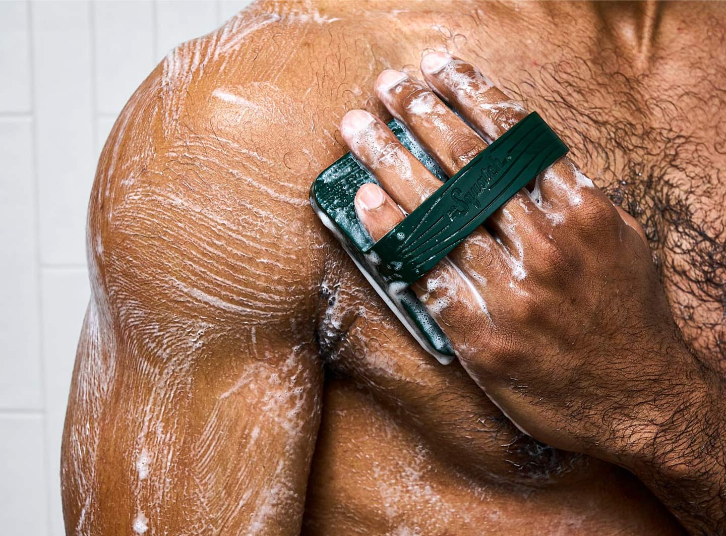 Person using a dark green soap scrubber on soapy shoulder in shower.