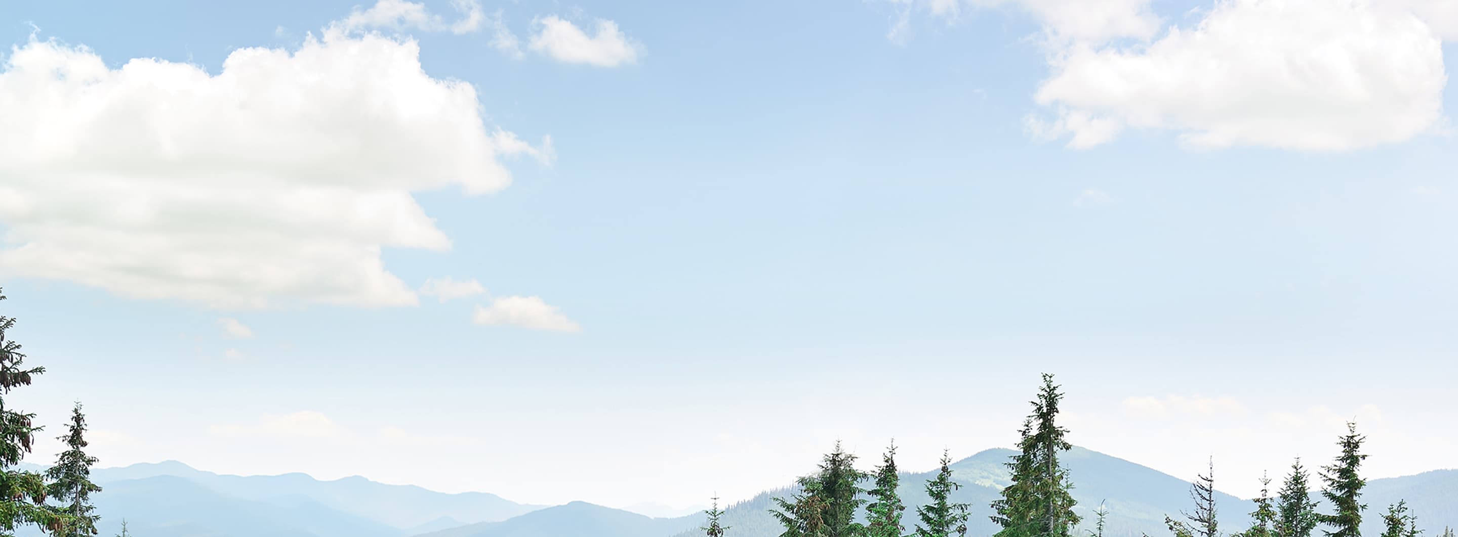 Clear sky above mountains and trees with fluffy clouds.