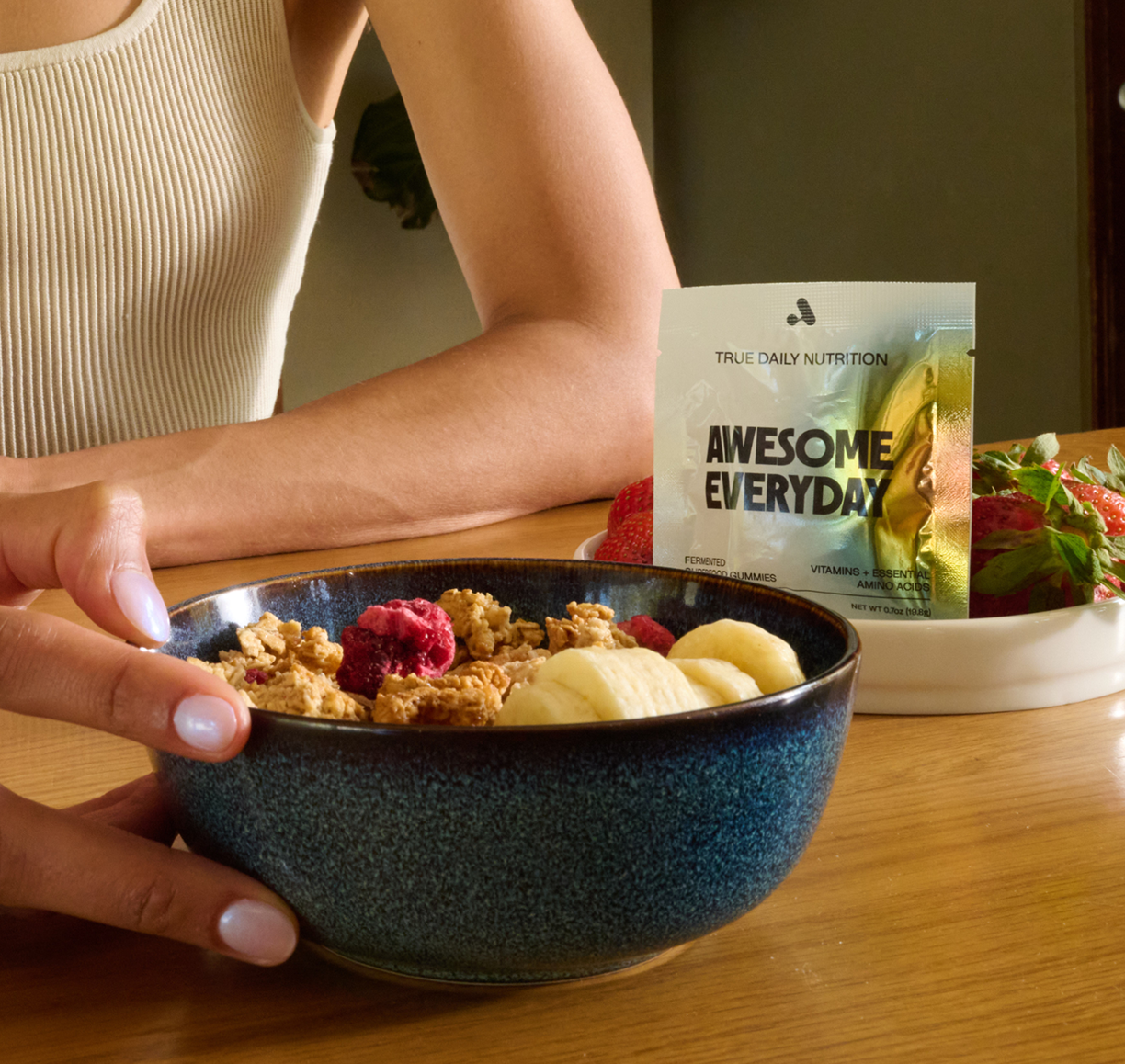 Person holding a bowl of cereal with banana and raspberries, next to a nutrition supplement pack.