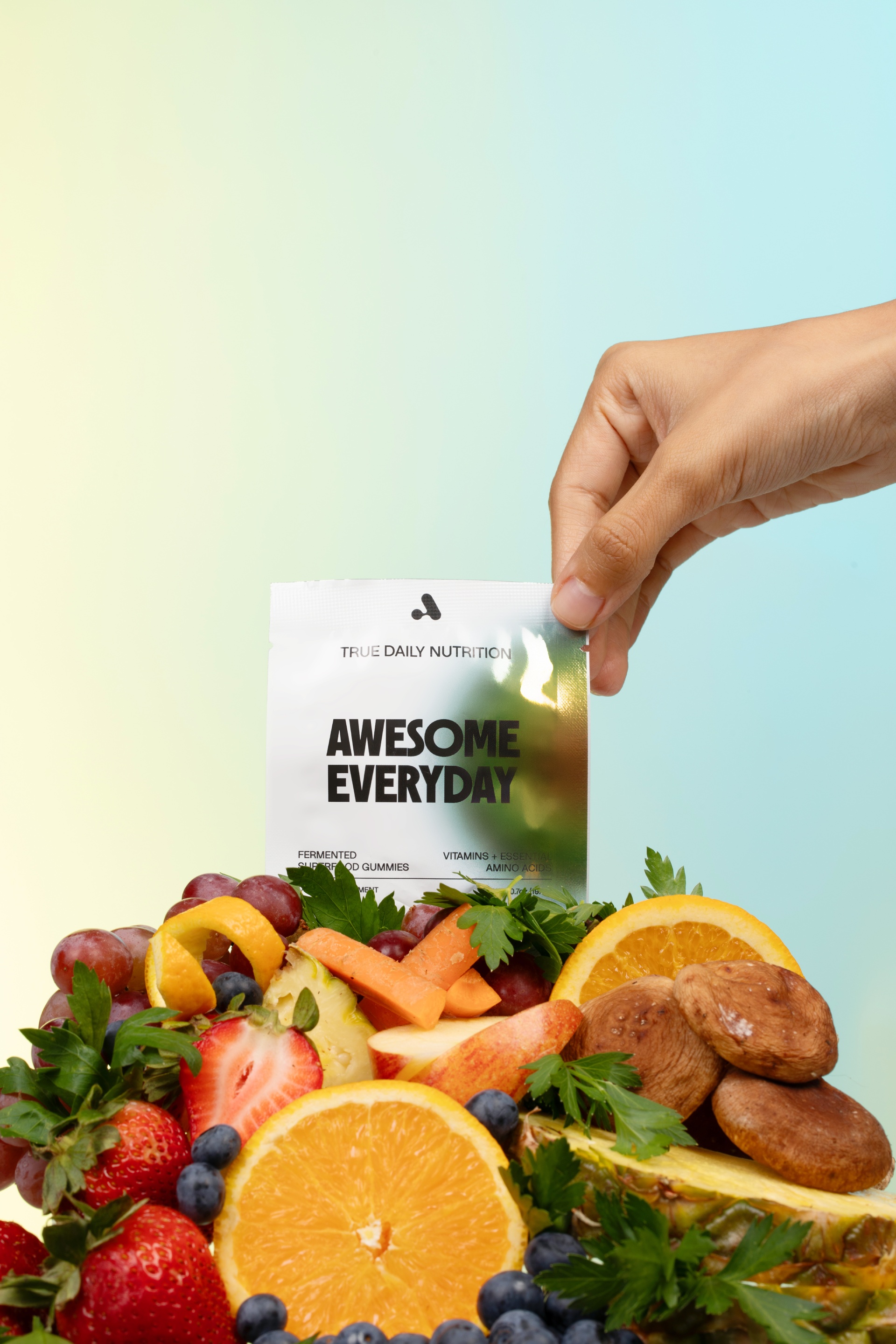 A hand holds a white packet of 'Awesome Everyday' supplements over an arrangement of fresh fruits and vegetables.