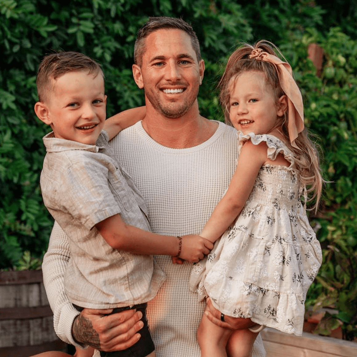 A person holding two children, smiling, with greenery in the background.