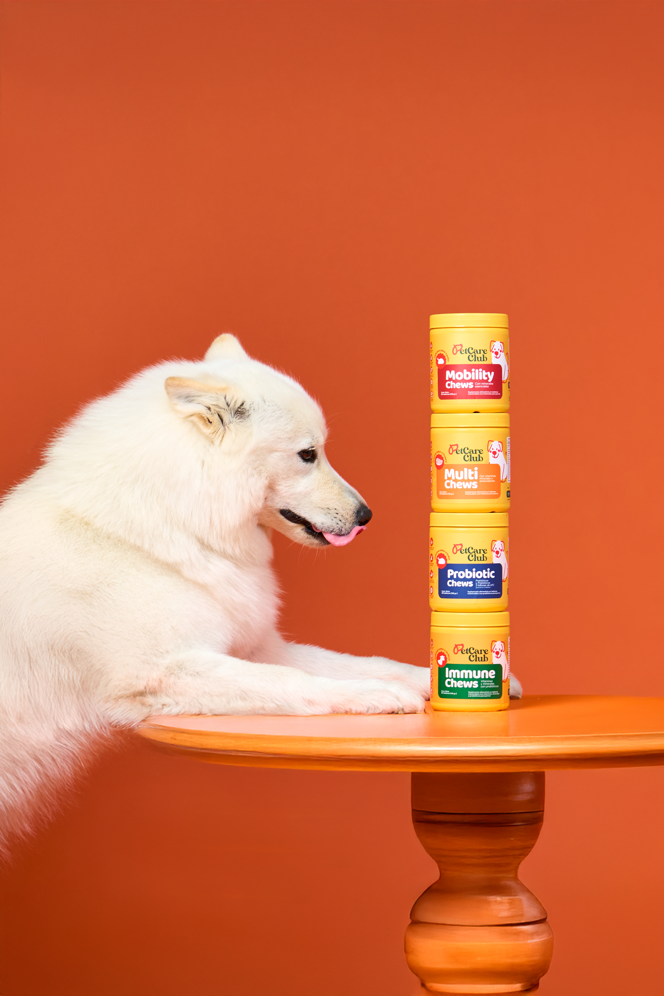 White dog with tongue out beside stacked pet supplement containers on orange table.