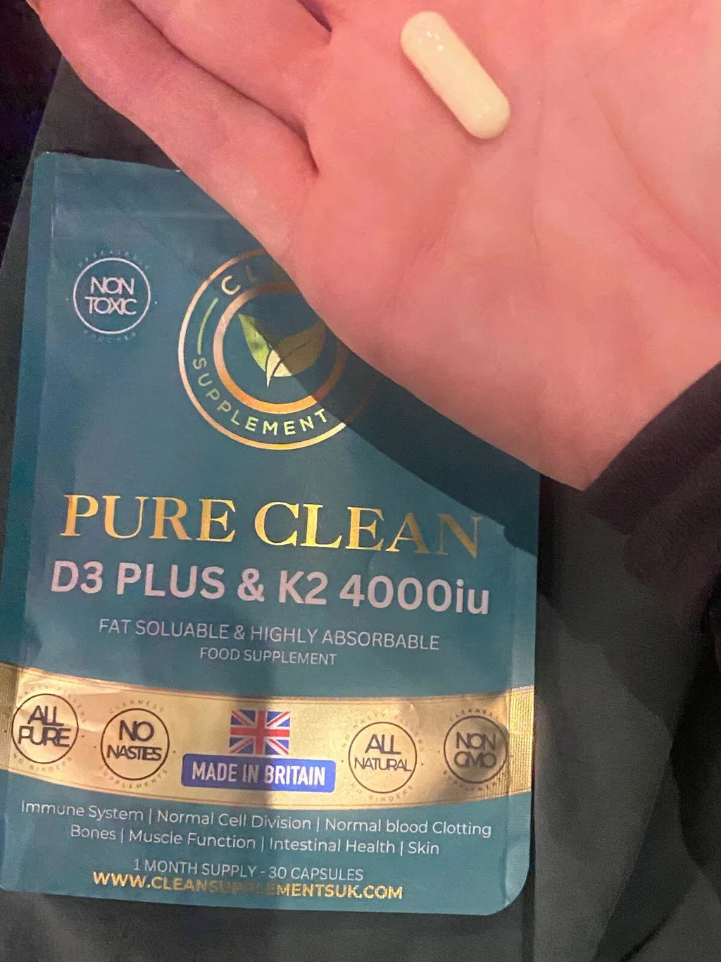 A hand holds a single capsule over a pouch of Pure Clean D3 Plus & K2 food supplement.