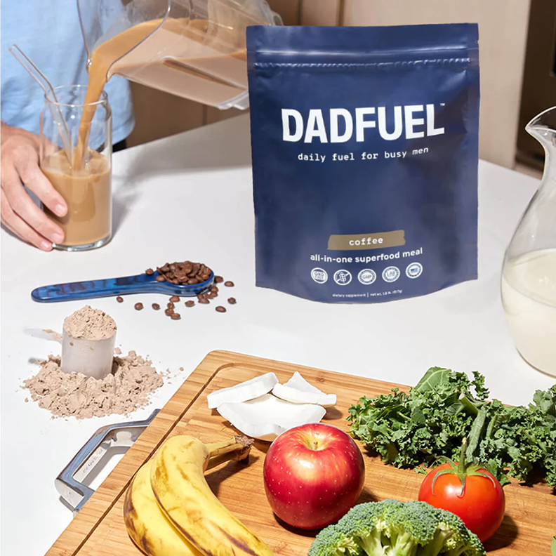 A bag of DADFUEL coffee meal powder next to fresh ingredients while a person pours a smoothie.