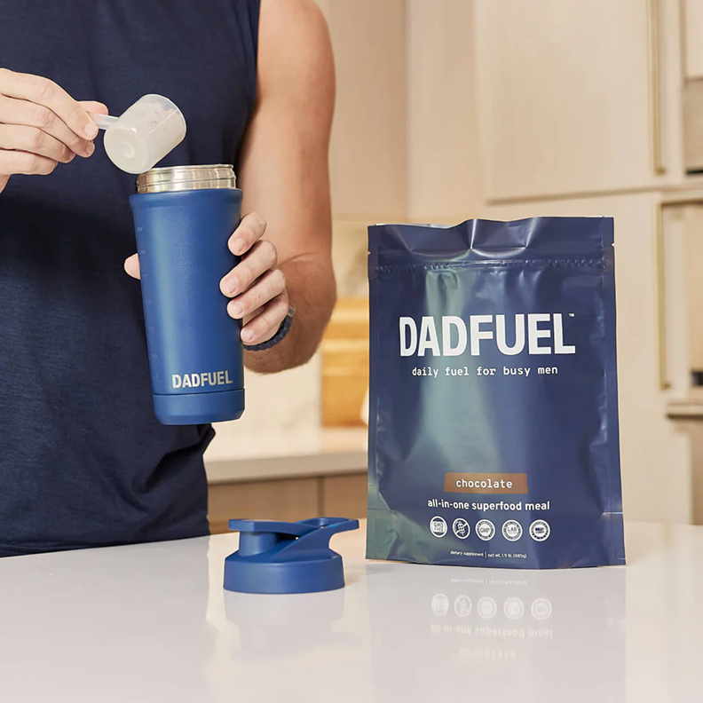 A man scoops Dadfuel chocolate powder into a blue shaker bottle next to the product bag.