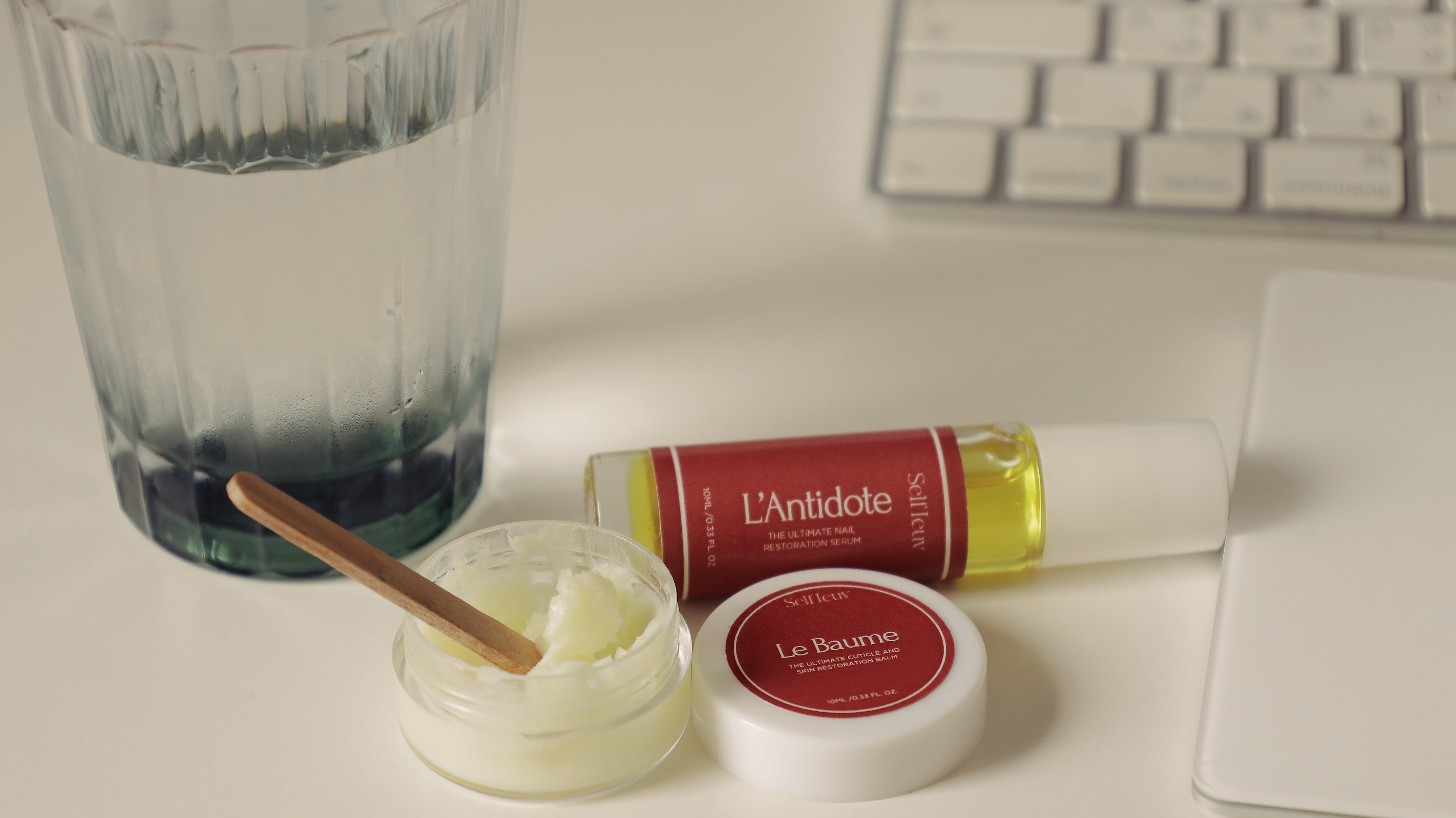 Glass of water, nail serum, balm, and keyboard on a desk.