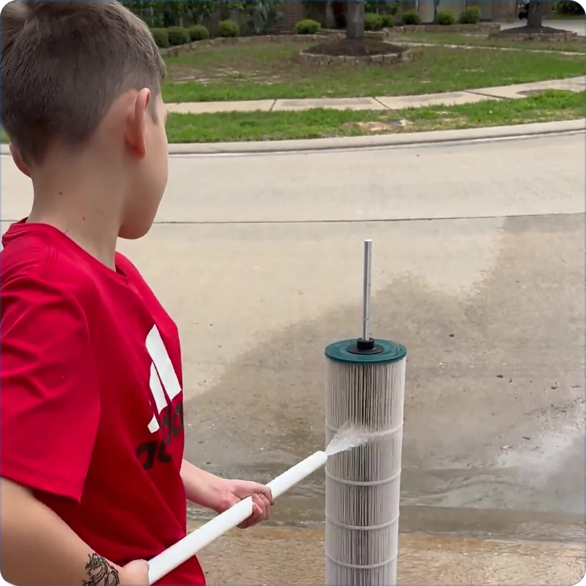 A boy in a red shirt sprays a large, cylindrical pleated filter with a hose on a driveway.