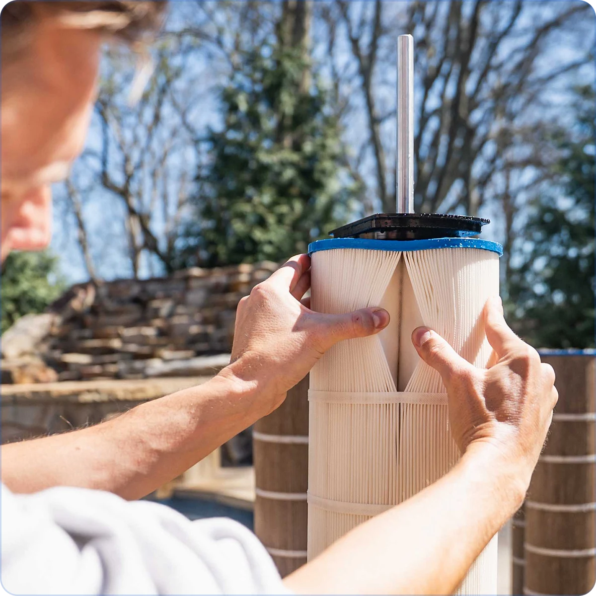 A person inspects the pleats of a large, cylindrical pool filter cartridge outdoors during the day.