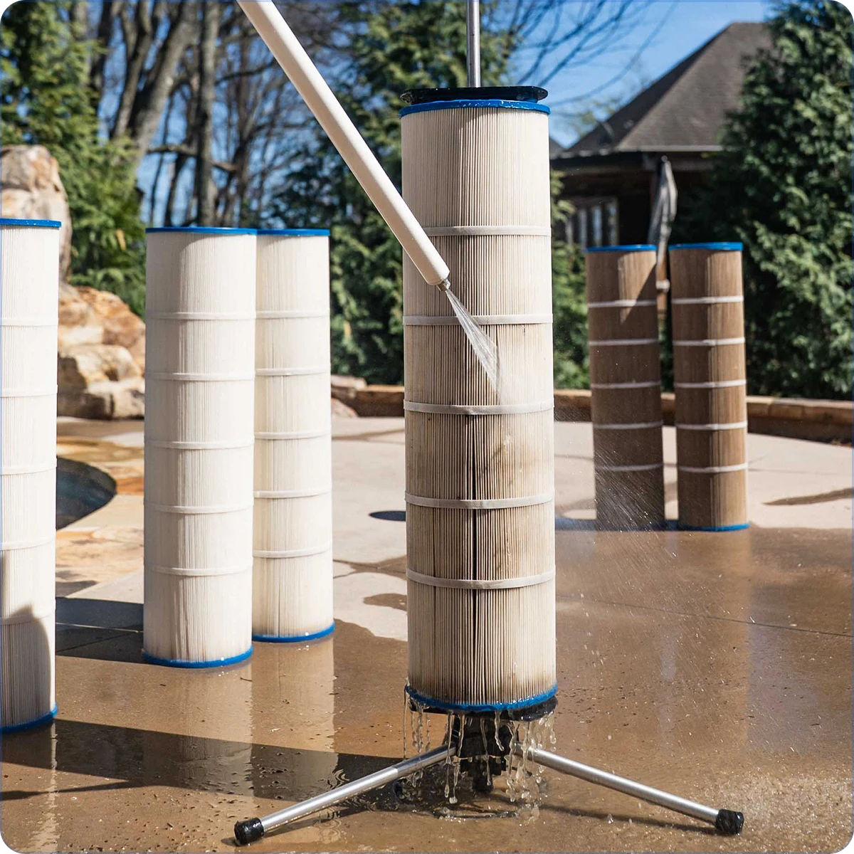 A person cleaning a dirty pool filter with a hose on an outdoor patio next to other filters.