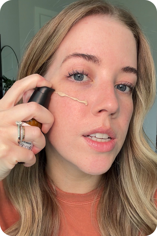 A woman applies liquid foundation from a pump bottle directly onto her cheek.