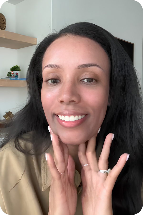 A close-up photo of a woman with dark hair smiling and touching her chin and neck.