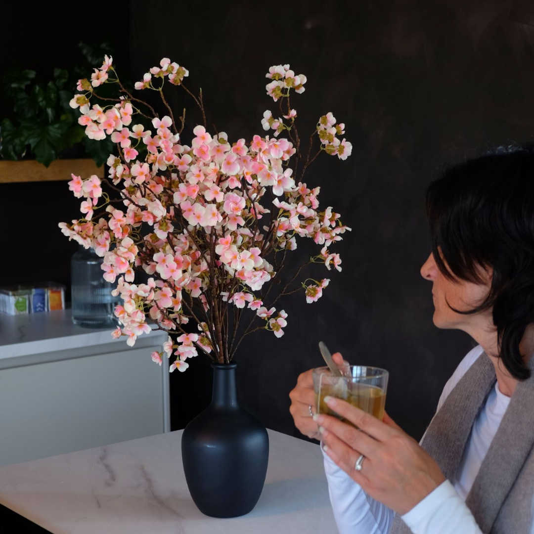 Person holding a cup of tea near a vase with pink flowers.